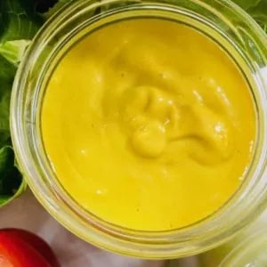 Gelbes Cheesy Dressing im Glas, umgeben von Salatblättern