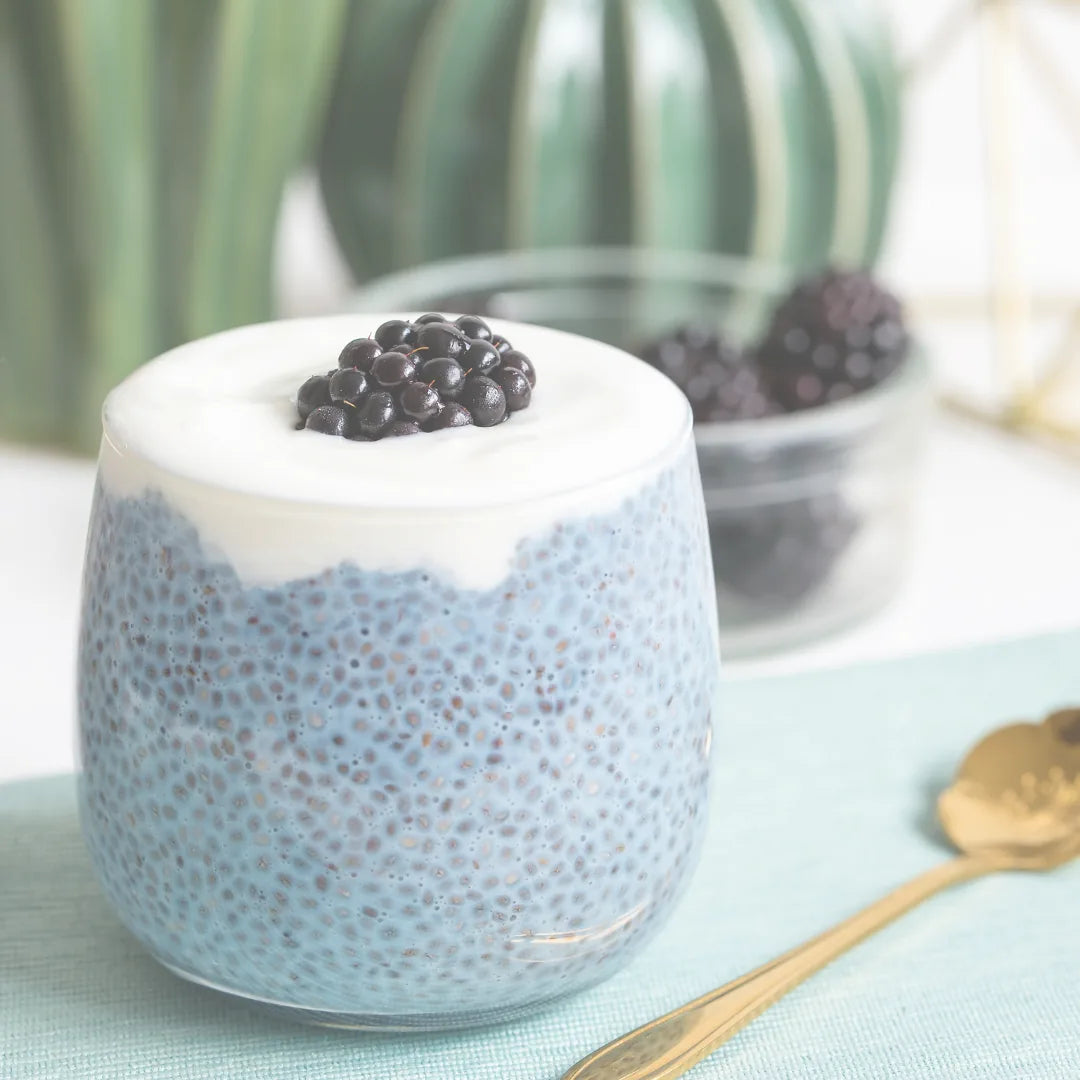 Blauer Chiapudding im Glas mit Creme und Brombeeren, daneben goldener Löffel